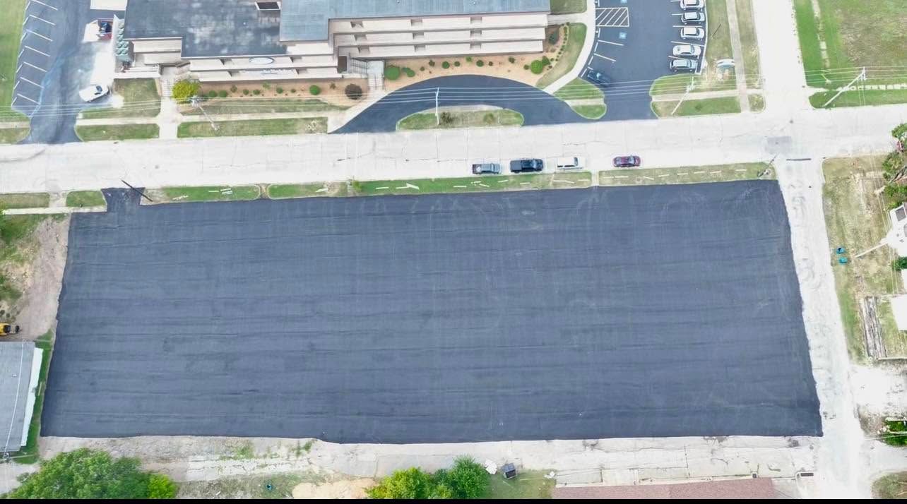 Aerial view of a large dark paved lot beside a street and buildings.