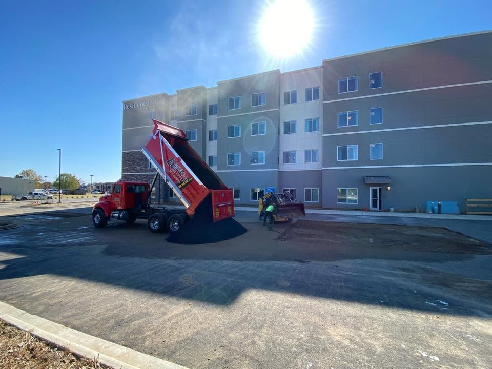 Red dump truck unloading gravel beside a gray apartment building on a sunny day