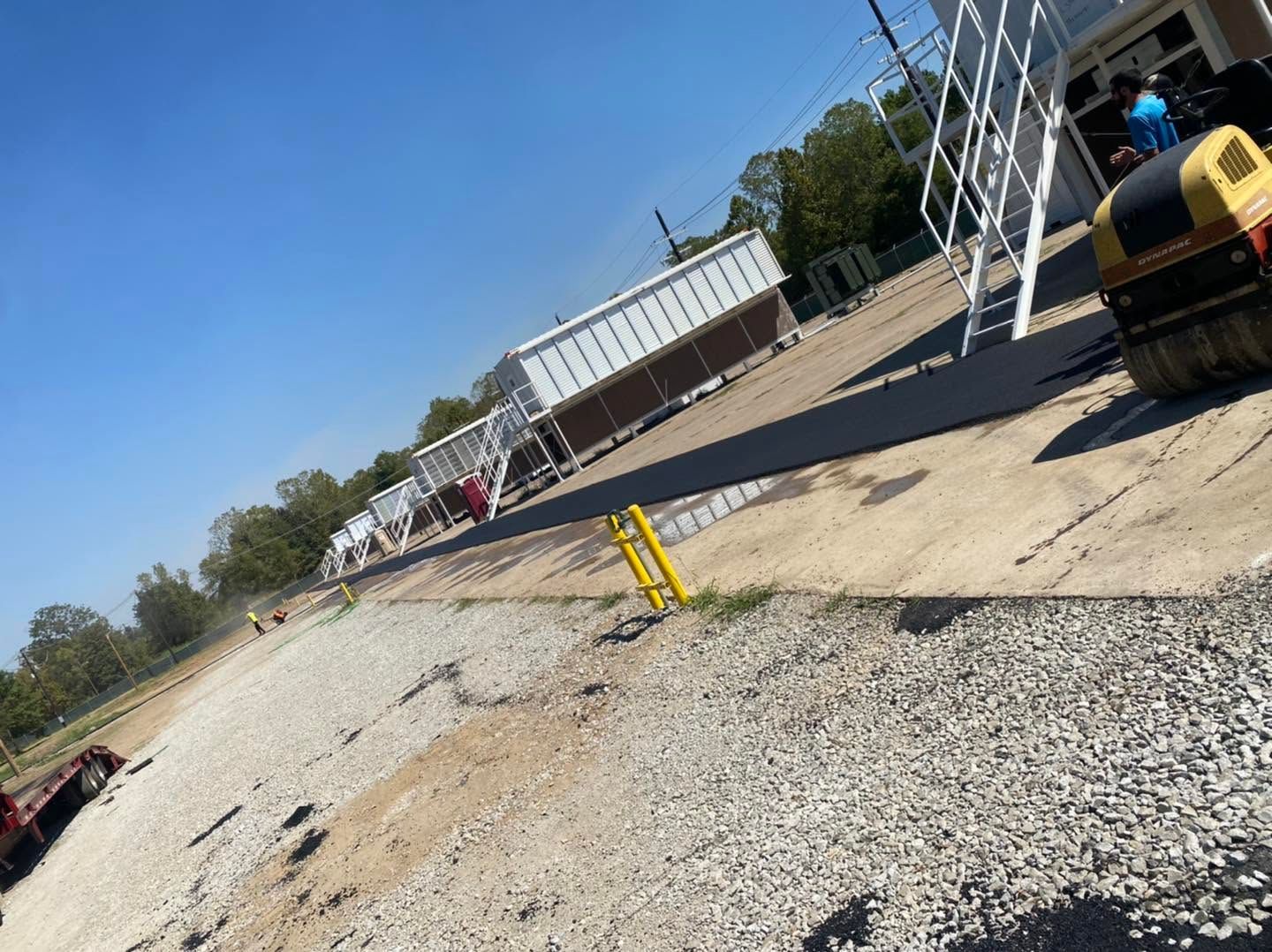 Dirt-covered industrial yard with a white building, gravel foreground, and a small yellow machine on the right