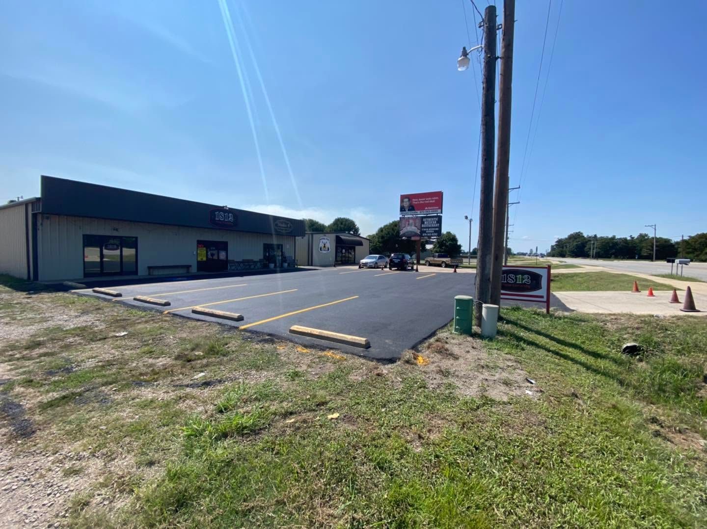 Small commercial building with empty parking lot, utility poles, and grassy roadside under a bright blue sky