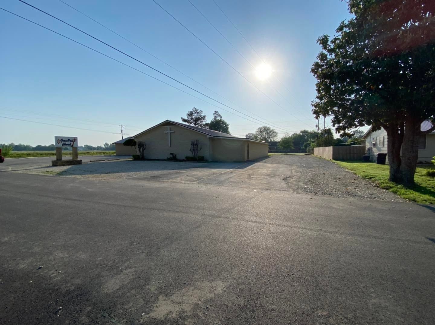 Sunlit rural parking lot beside a low beige building and large tree under a clear blue sky