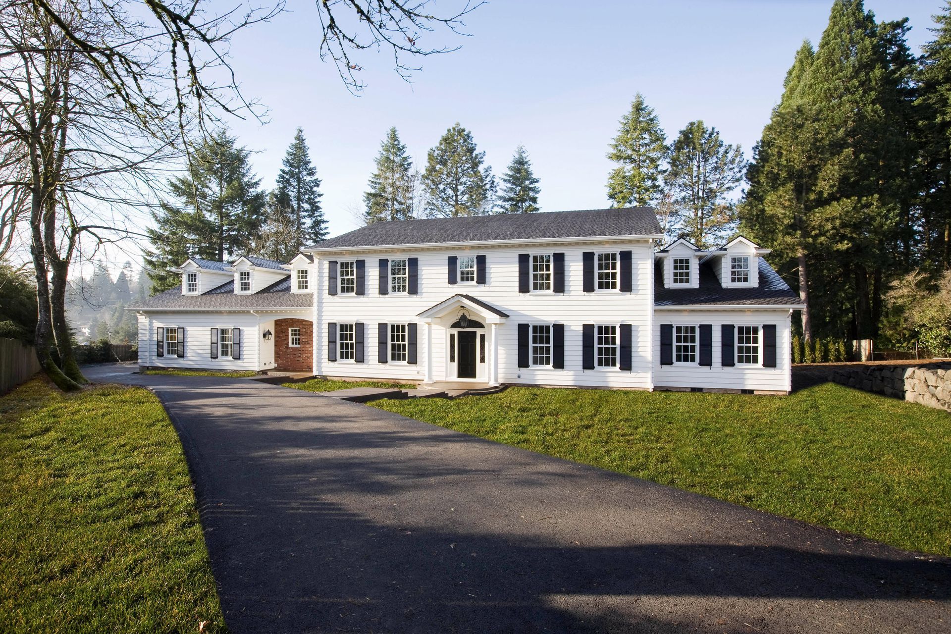 Large white colonial house with black shutters and a paved driveway, surrounded by trees and lawn