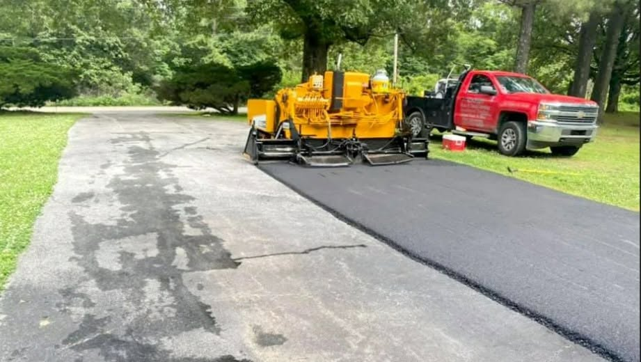 Red truck and yellow paving machine laying fresh black asphalt on a driveway beneath trees