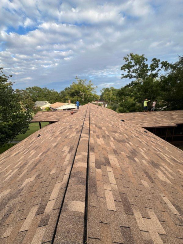 Roof with brown shingles, blue sky, trees in the background. People working.