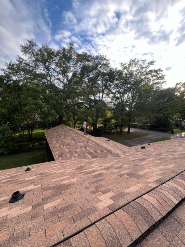 Roof with new shingles, a person standing on it, trees in the background, blue sky with clouds.