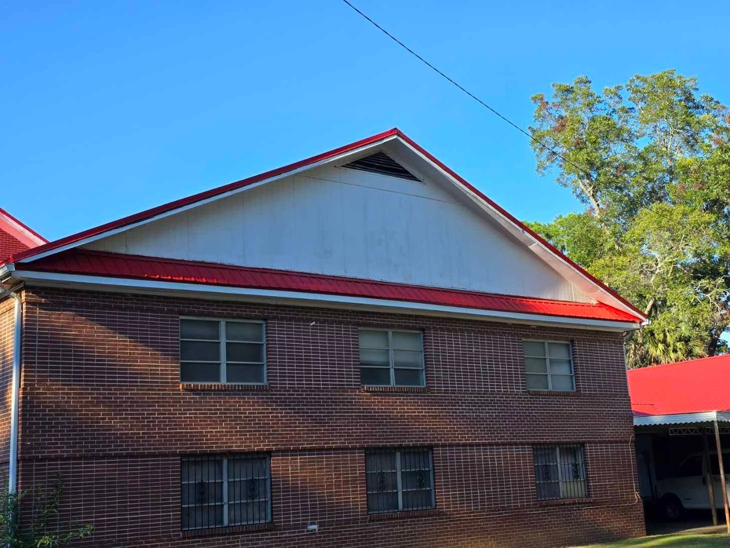 Two-story brick building with red roof against a blue sky, trees in background.