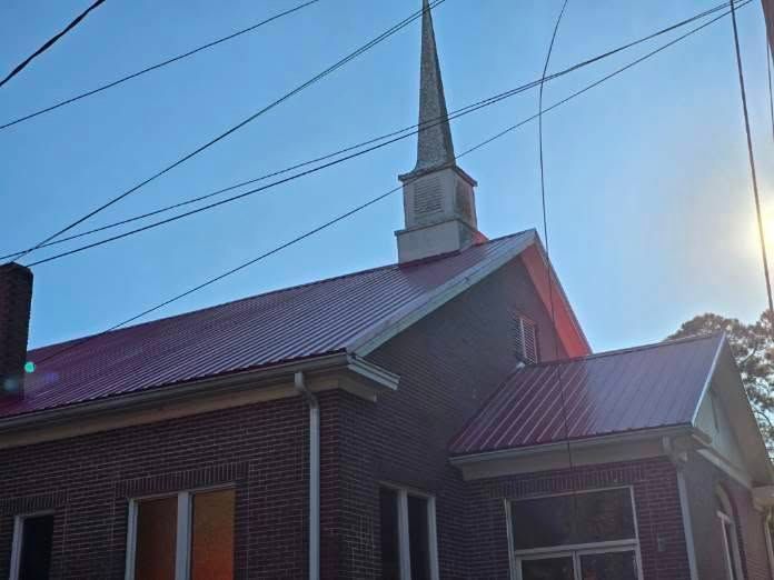Red-roofed brick church with steeple and power lines against a blue sky.