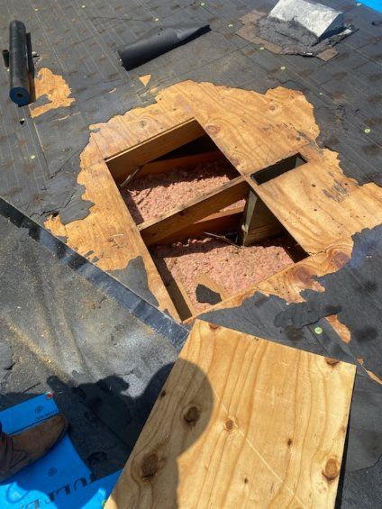 Rooftop repair: hole cut in plywood, revealing insulation. Black roofing material and tools visible.