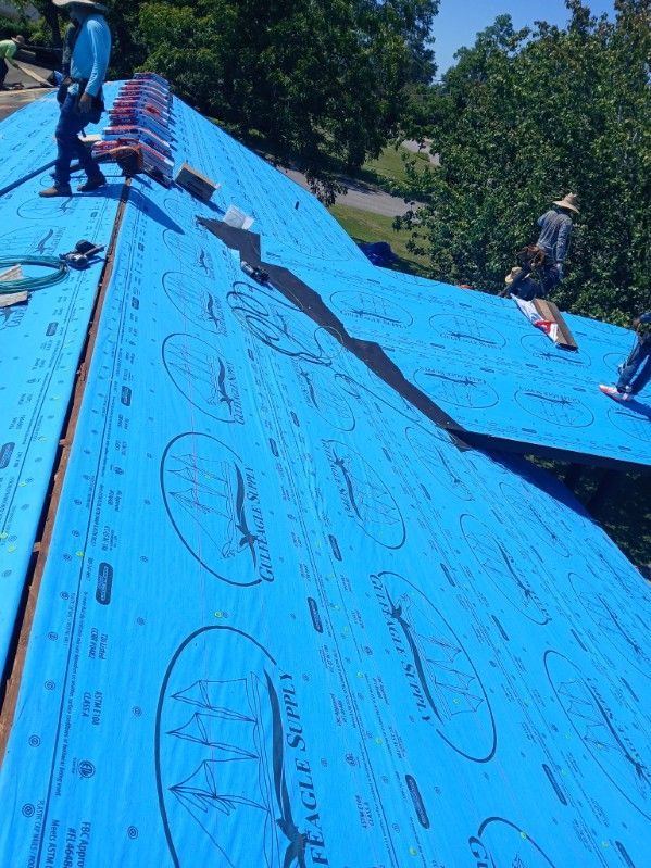 Roofers install blue underlayment on a rooftop under a bright sky.
