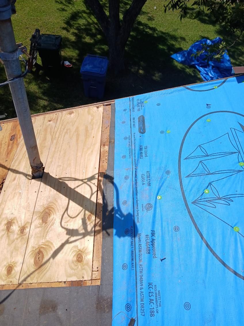 Roofing work in progress, showing plywood and blue underlayment, with tools and a tree in the background.