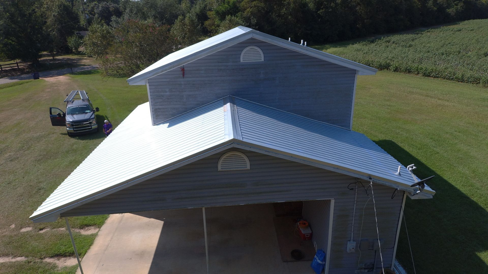 Grey metal-roofed building with carport, two vehicles parked nearby, grassy field.