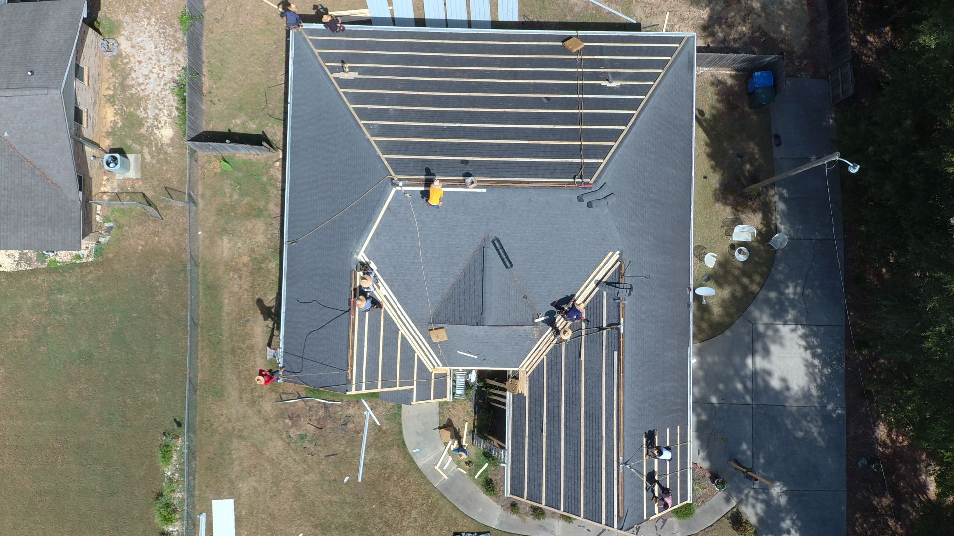 Overhead view of a house roof under construction. Workers on roof, black shingles, wood beams visible.