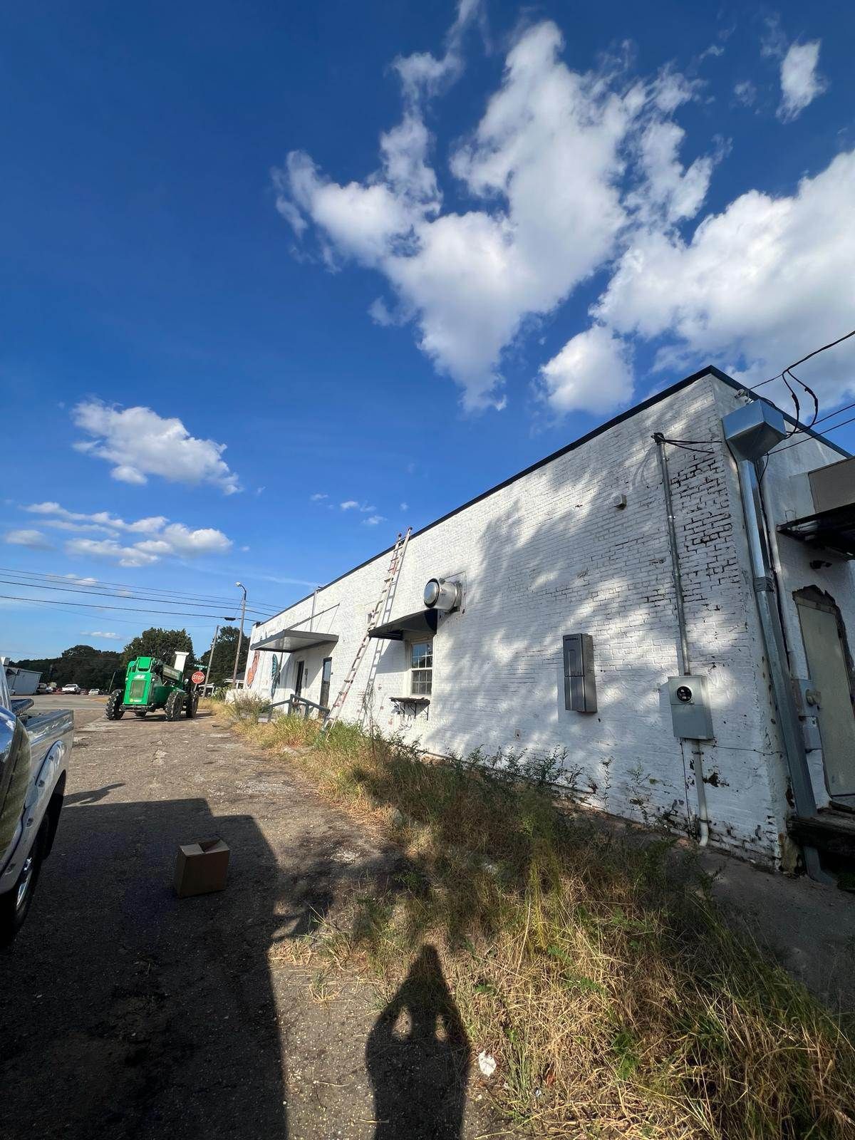 White brick building with overgrown weeds. Blue sky with clouds. Green lift in the background.