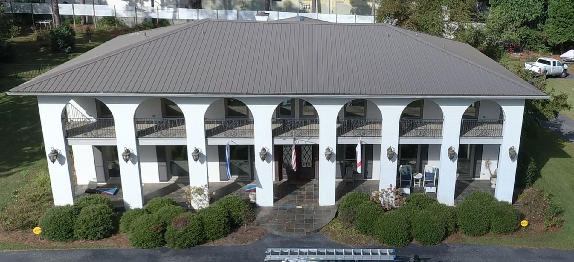 White building with arched facade and gray metal roof, surrounded by greenery.