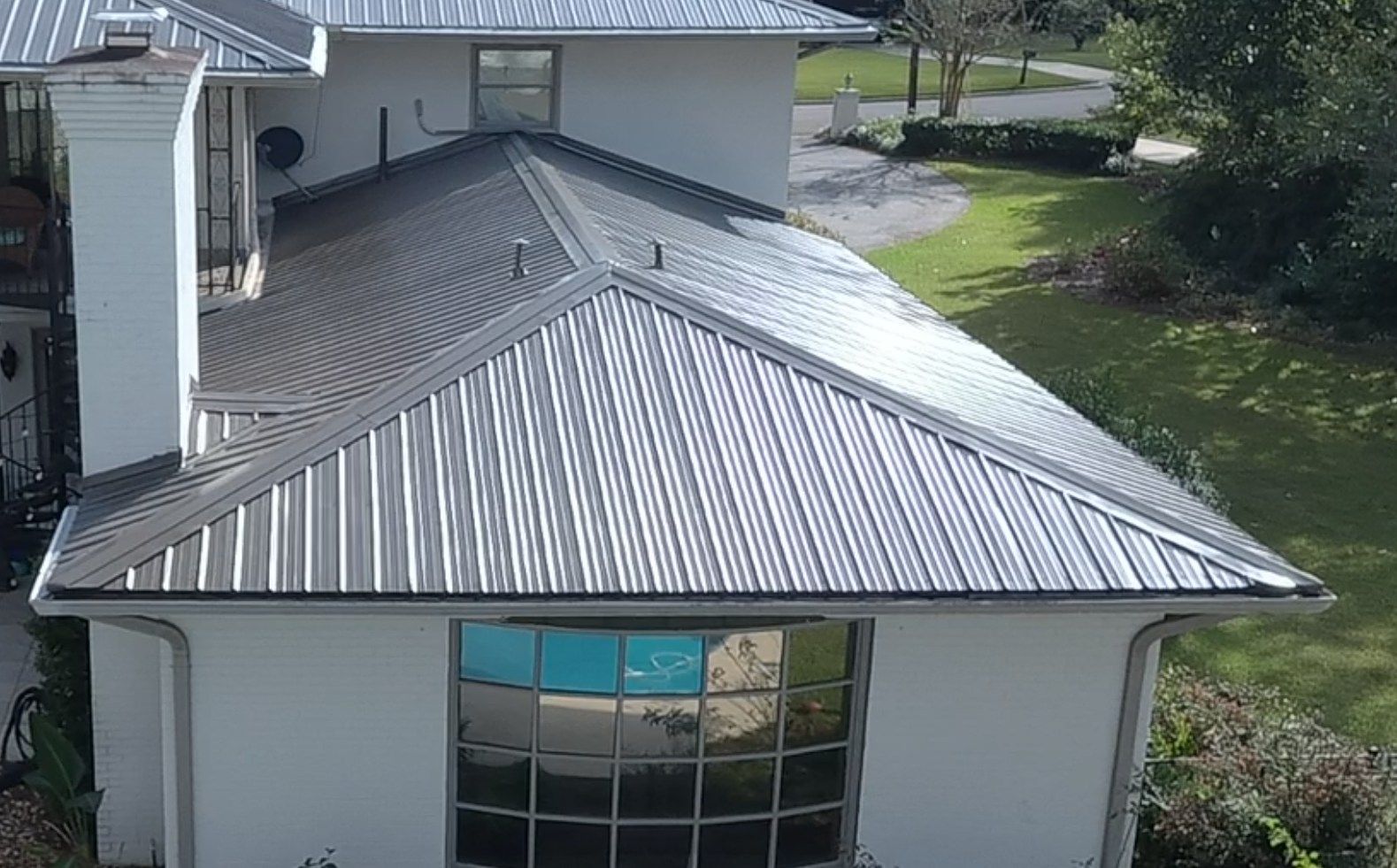 A house with a silver metal roof reflects a swimming pool. Sunlight on green grass nearby.