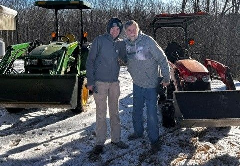 Two people standing between green and red tractors in a snowy field, smiling at the camera