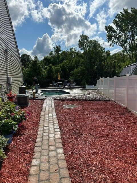 Backyard walkway with red mulch, white fence, and a pool under a cloudy sky