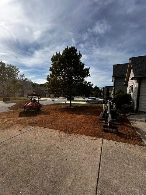 Front yard with a large tree, fresh mulch, a wheelbarrow, and a house under a cloudy sky