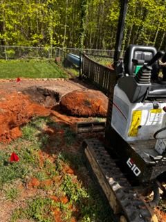 Excavator digging near a large tree stump in a grassy yard beside a fence