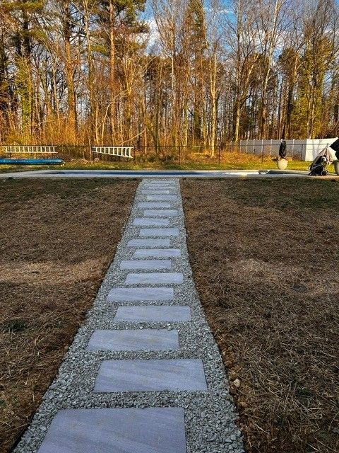 Stone path leading through a grassy yard toward trees in autumn, with a bench and small structure in the background