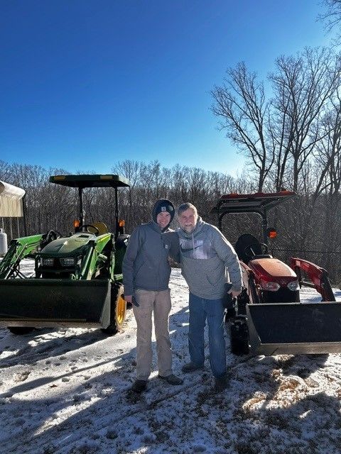 Two people standing between green and red tractors in a snowy field under a clear blue sky