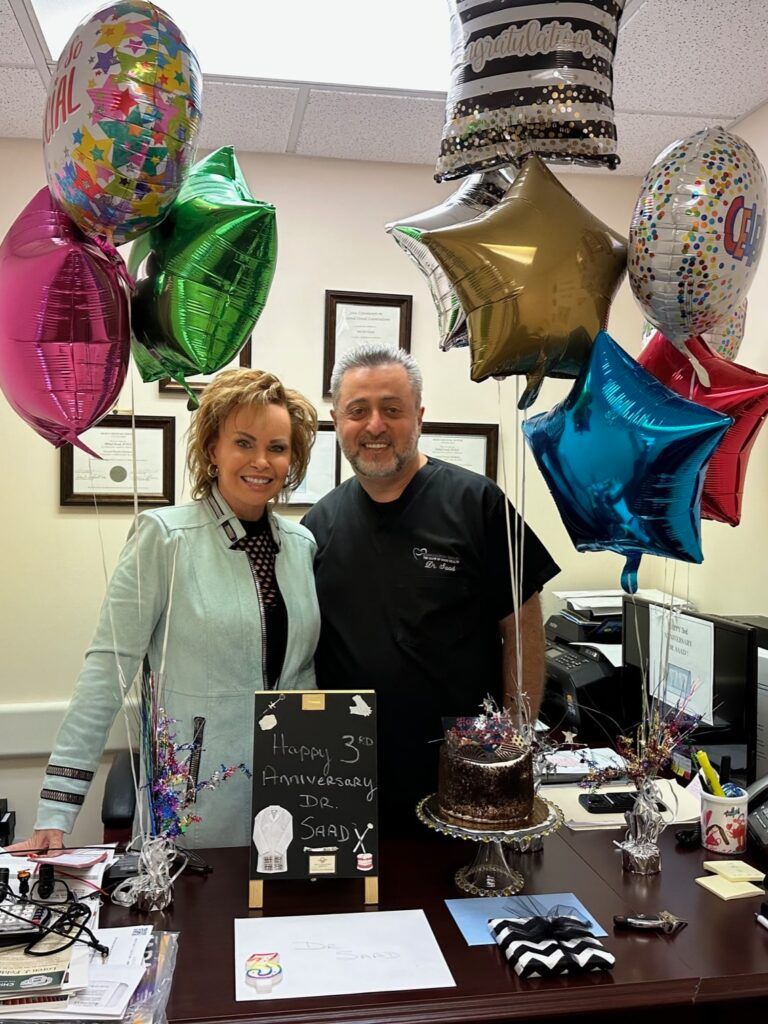 A man and a woman are standing in front of balloons and a cake.