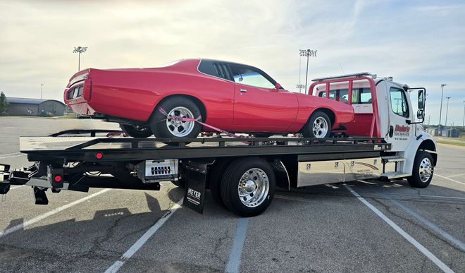 Red car on a tow truck on a highway under a clear blue sky.