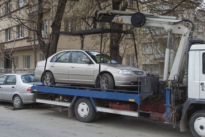 Tow truck loading a silver sedan on a blue flatbed in front of apartment buildings.