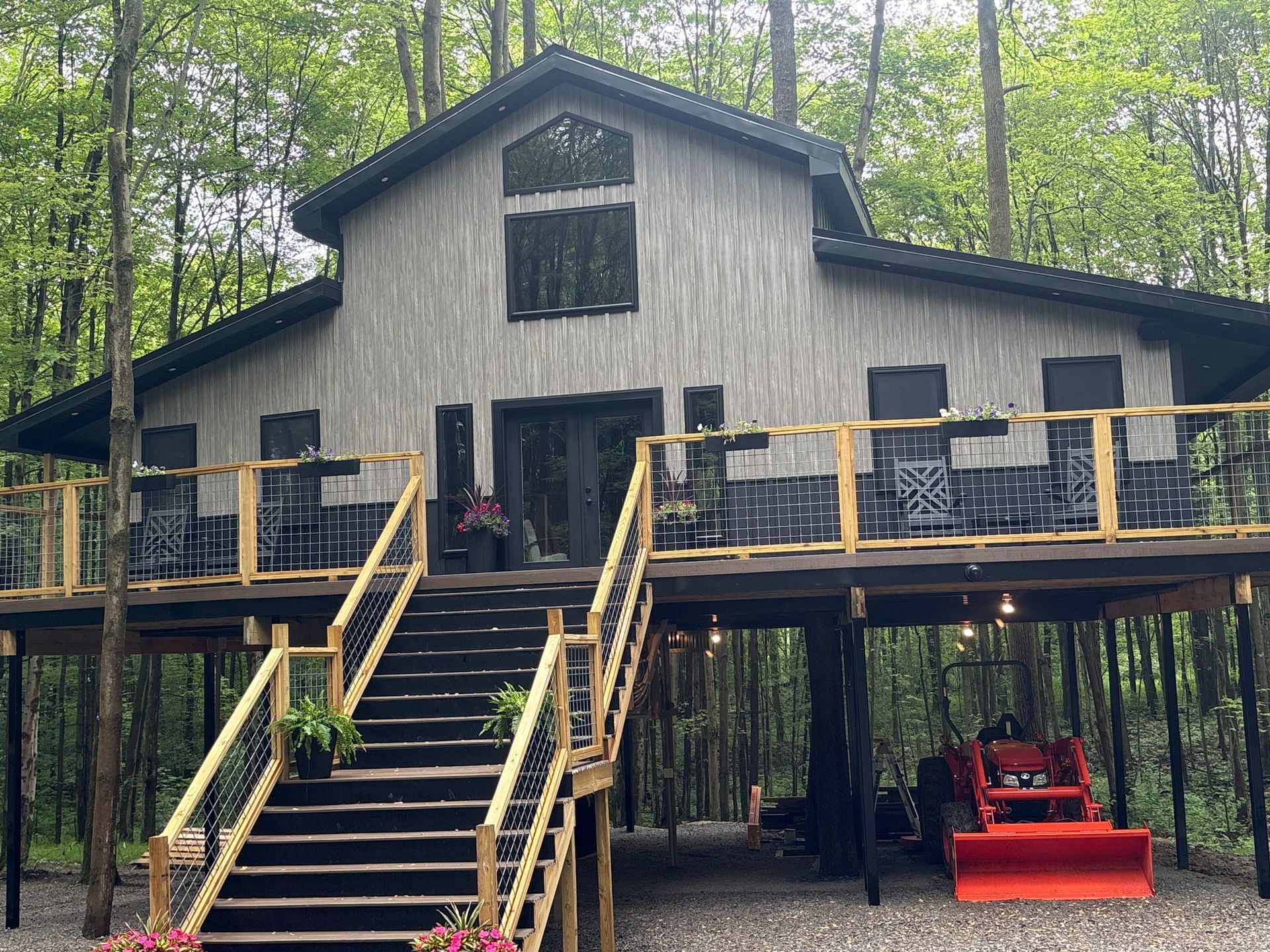 Rustic cabin with a wide staircase and deck in a wooded setting, with a red tractor below.