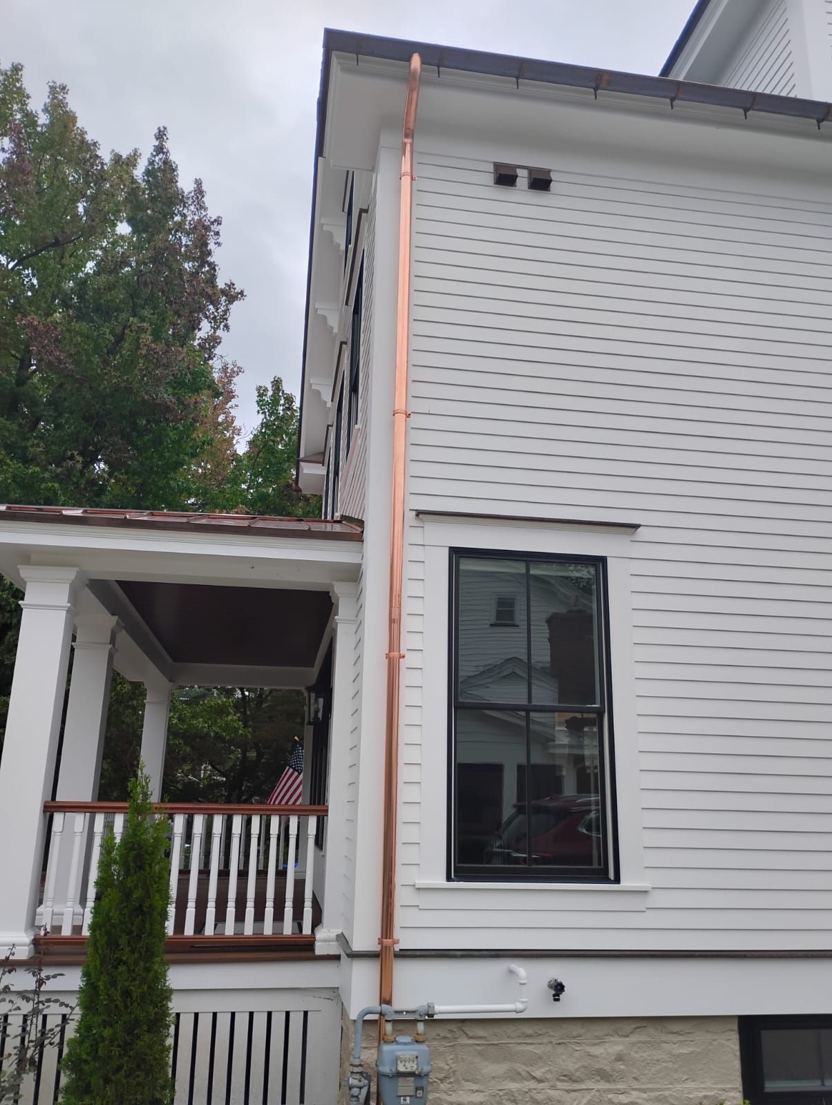 White house exterior with porch, black-framed window, and orange downspout beside a tall evergreen tree