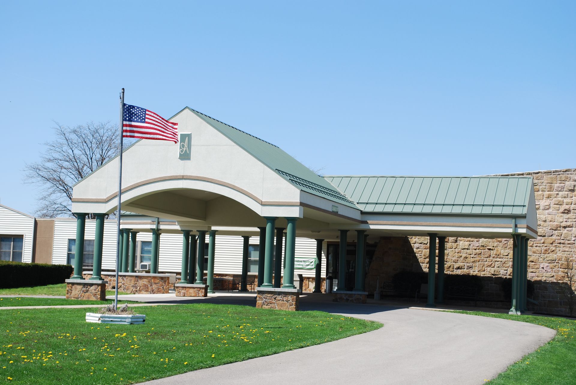 Covered building entrance with American flag, green roof, and curved driveway on a sunny day