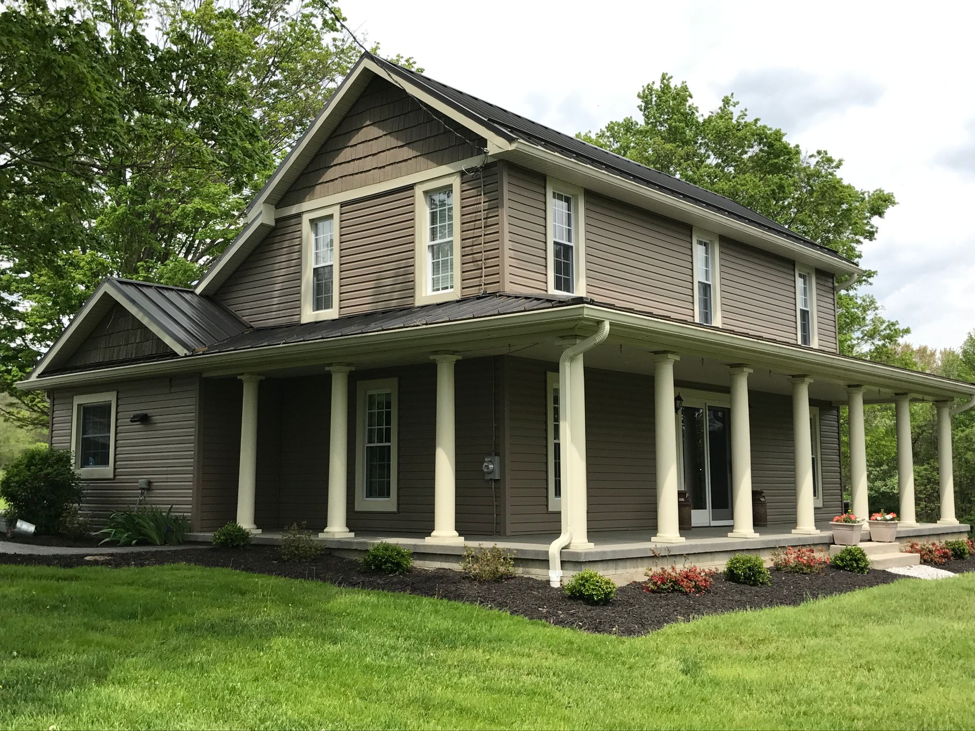 Two-story house with a wraparound porch and white columns, surrounded by green lawn and trees.