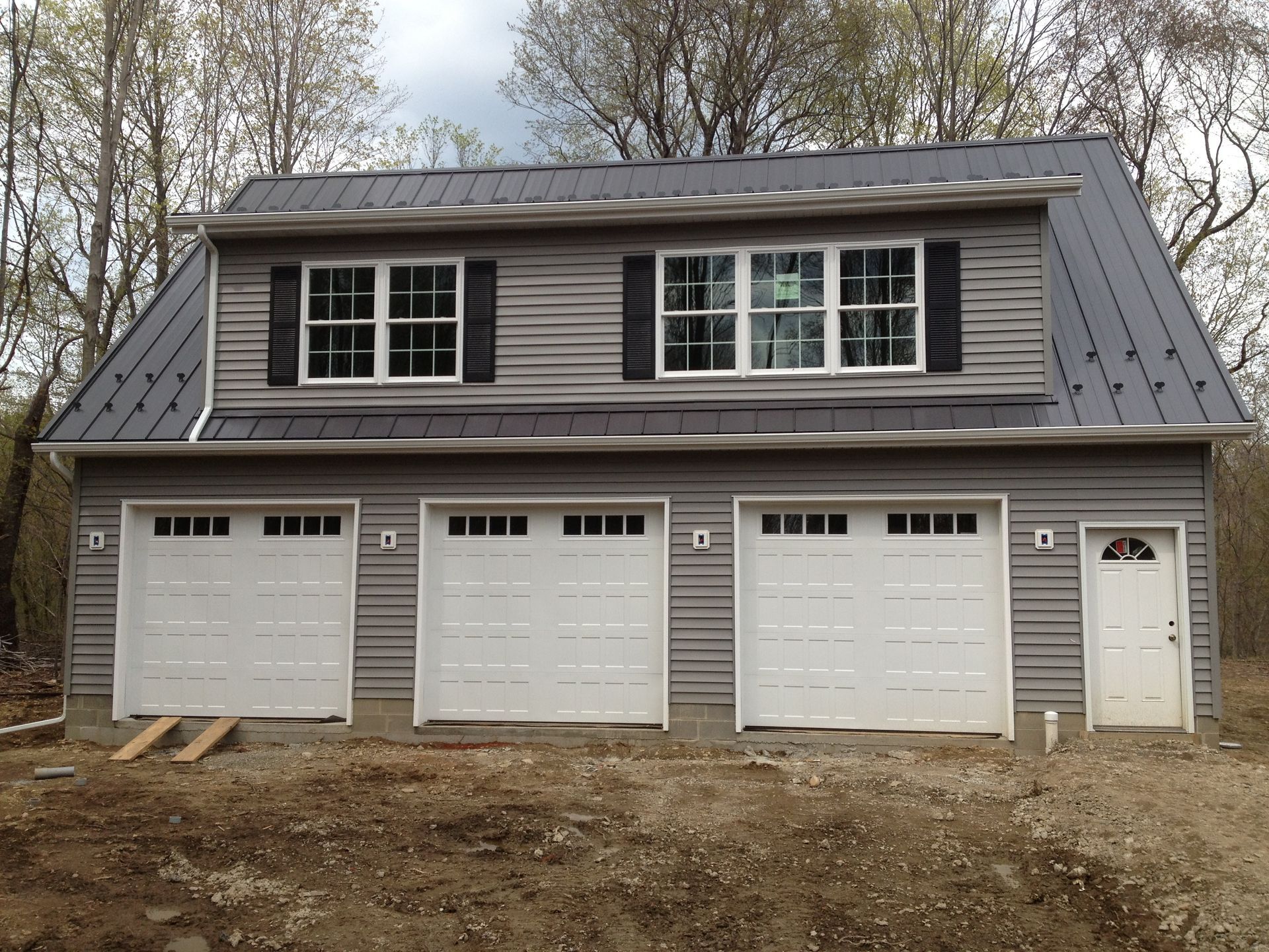 Gray garage with three doors and a white side entrance, beneath a dormer on a wooded lot