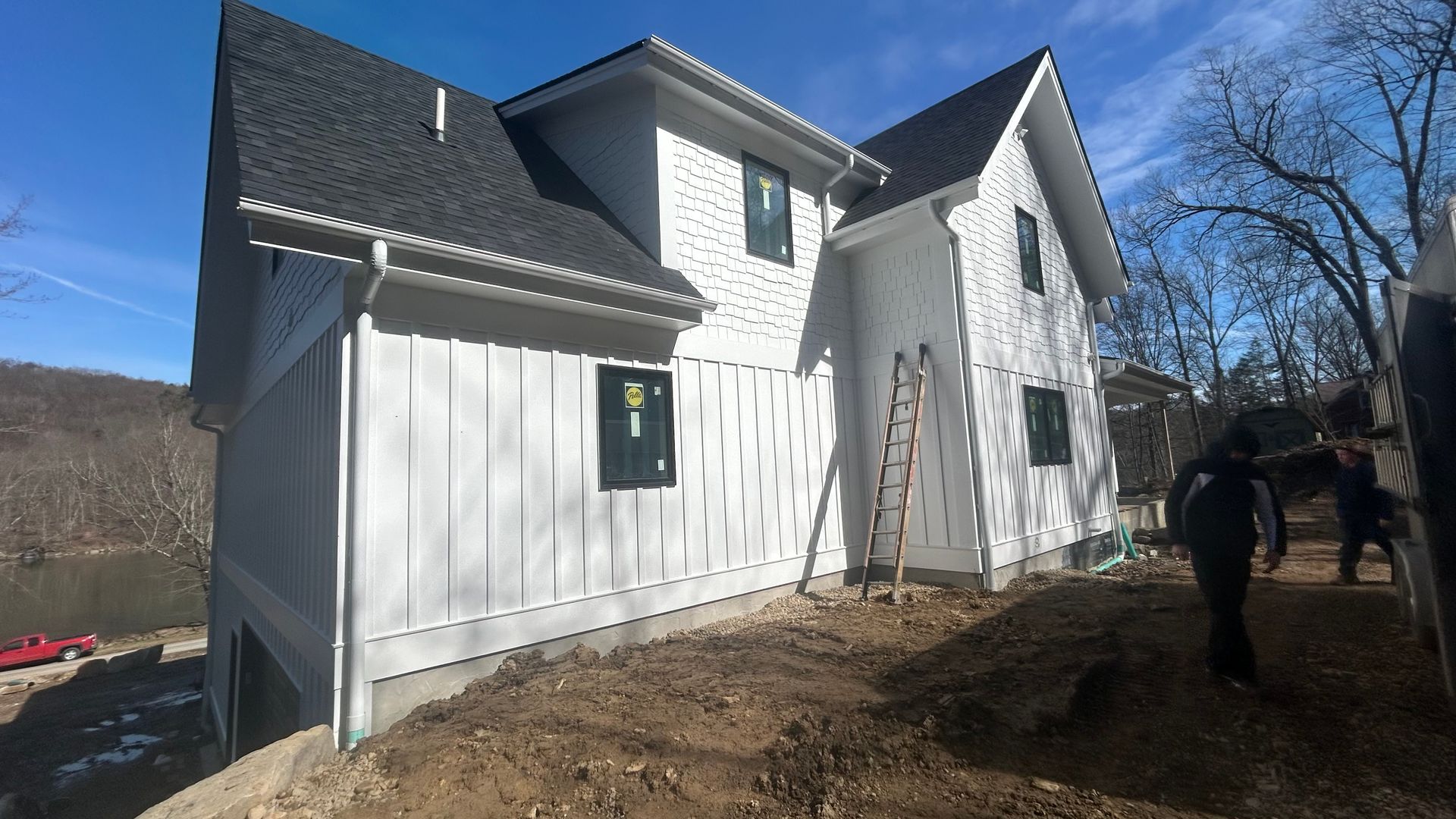 White two-story house under construction beside bare trees, with a person standing near the right side.