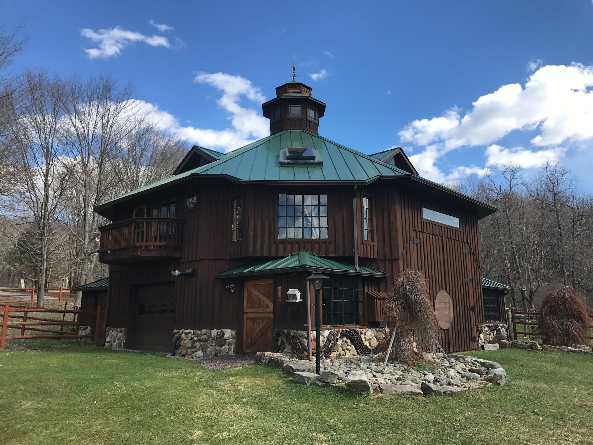 Round wooden house with teal roof and porch in a grassy yard under a blue sky with clouds