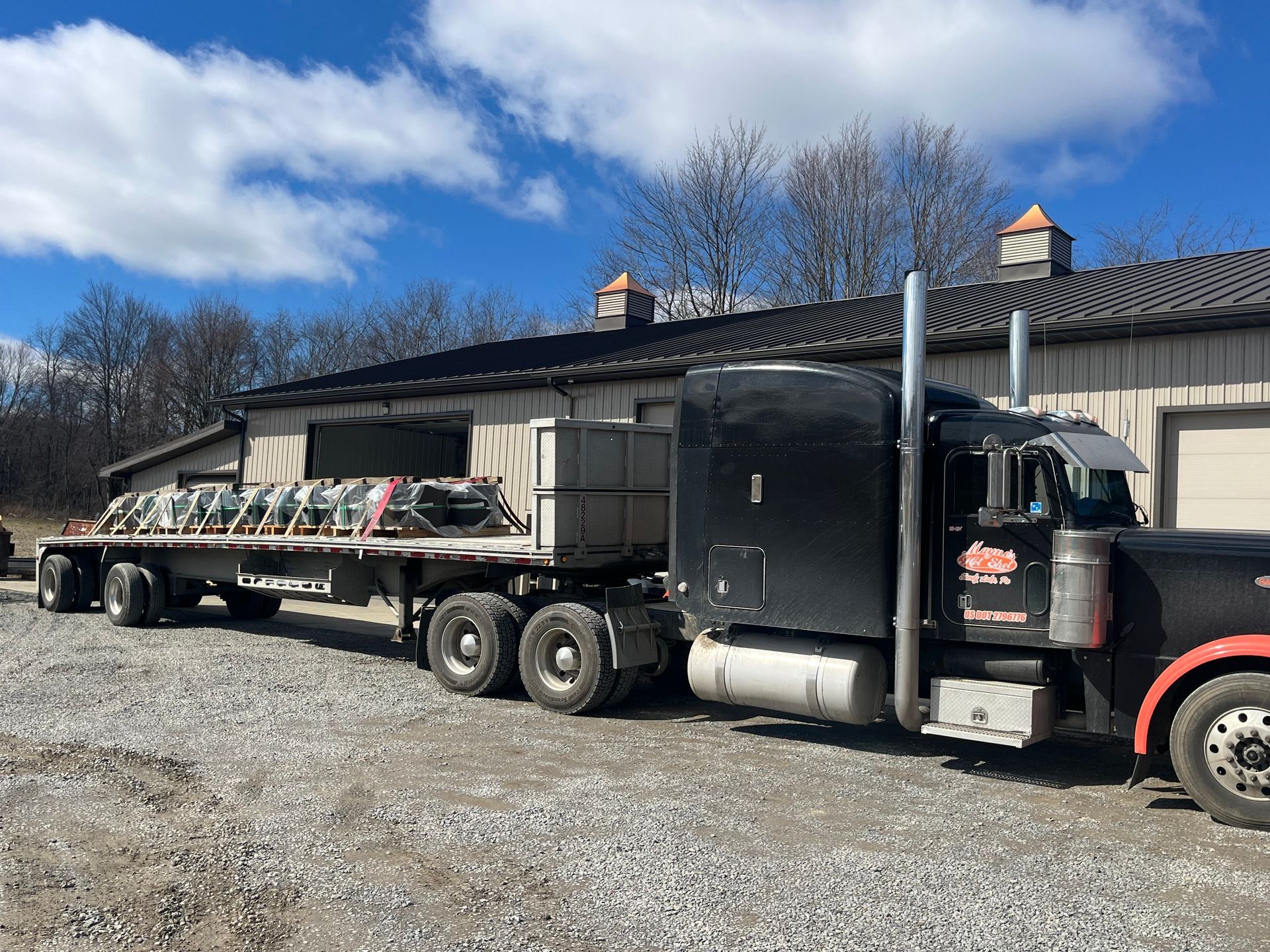 Black semi-truck hauling a long flatbed of stone slabs beside a metal building under a blue sky