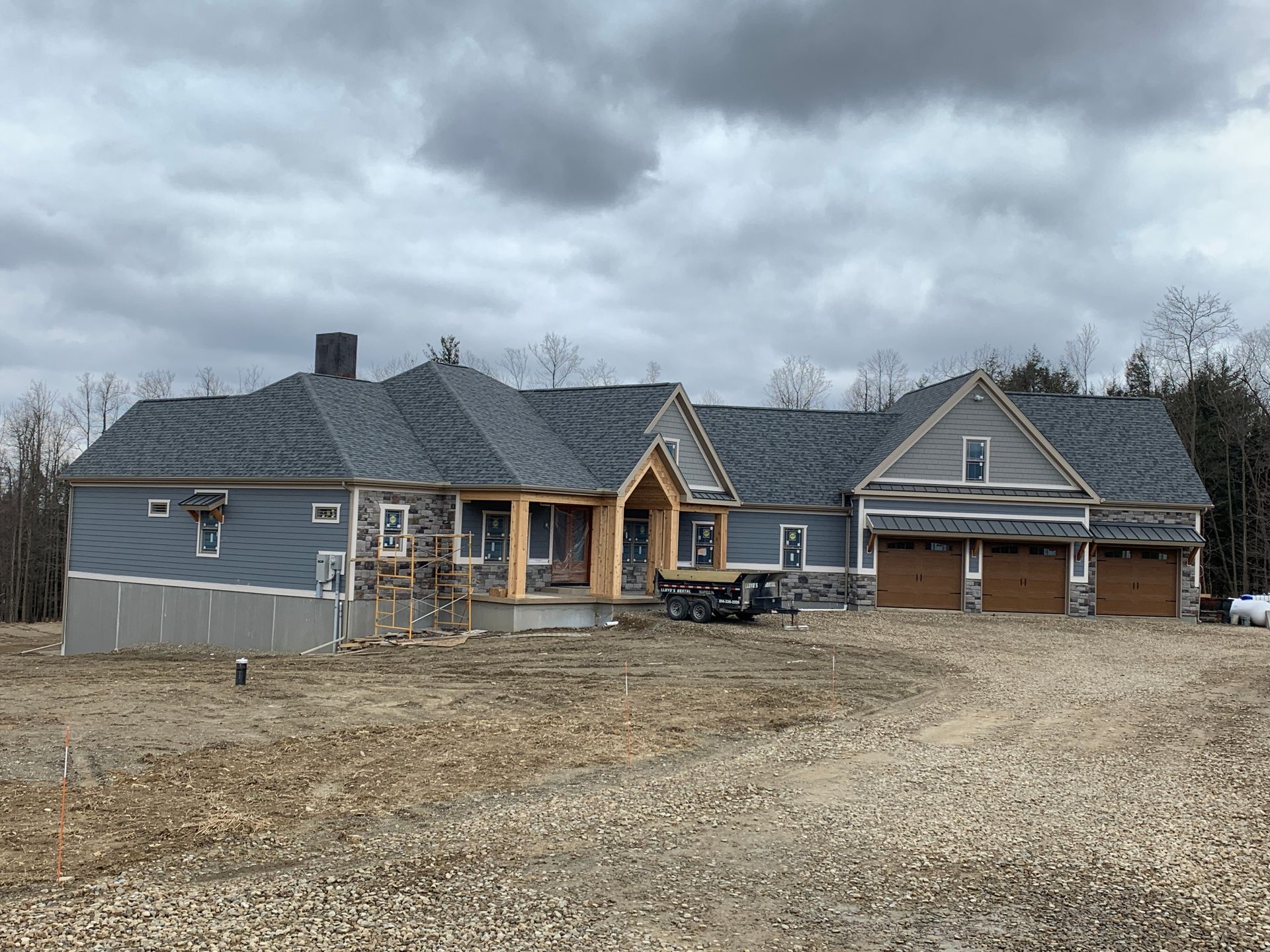 Large unfinished house with gray siding, wood framing, and a gravel driveway under cloudy skies