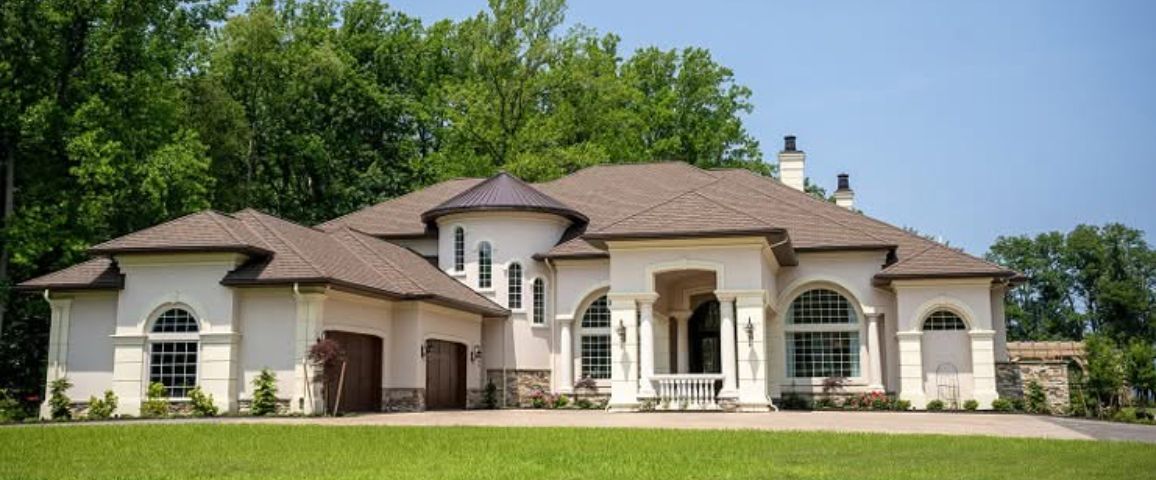 Large beige suburban house with a red-tiled roof, arched windows, and a manicured lawn under a blue sky