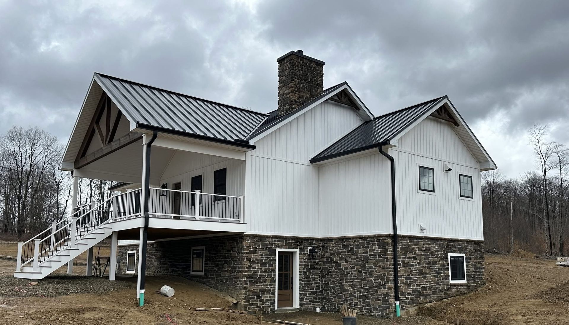 White house with black metal roof and stone foundation, viewed from the side in a wooded rural setting