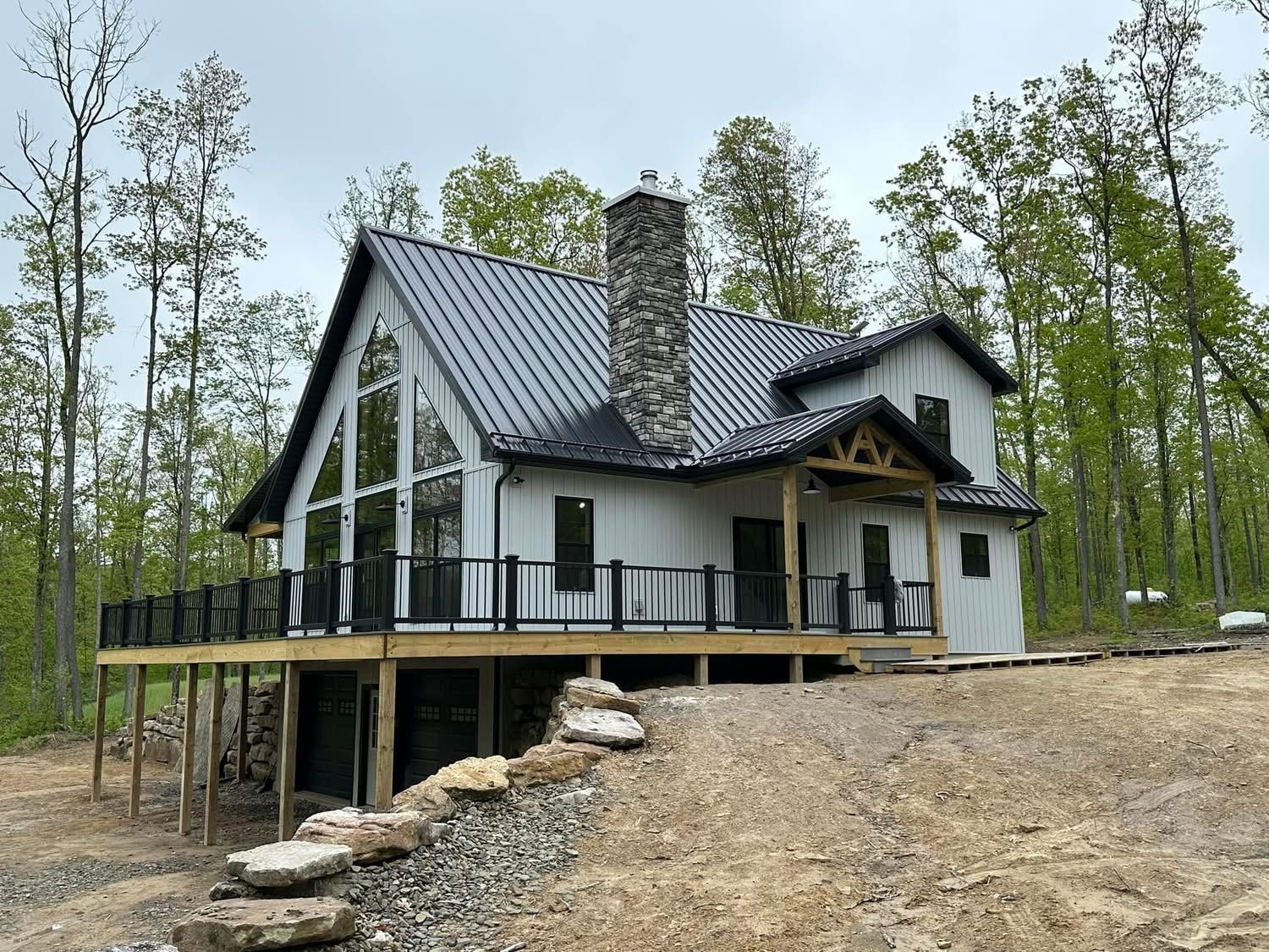 Modern white house with dark metal roof and stone chimney on a raised deck in a wooded clearing