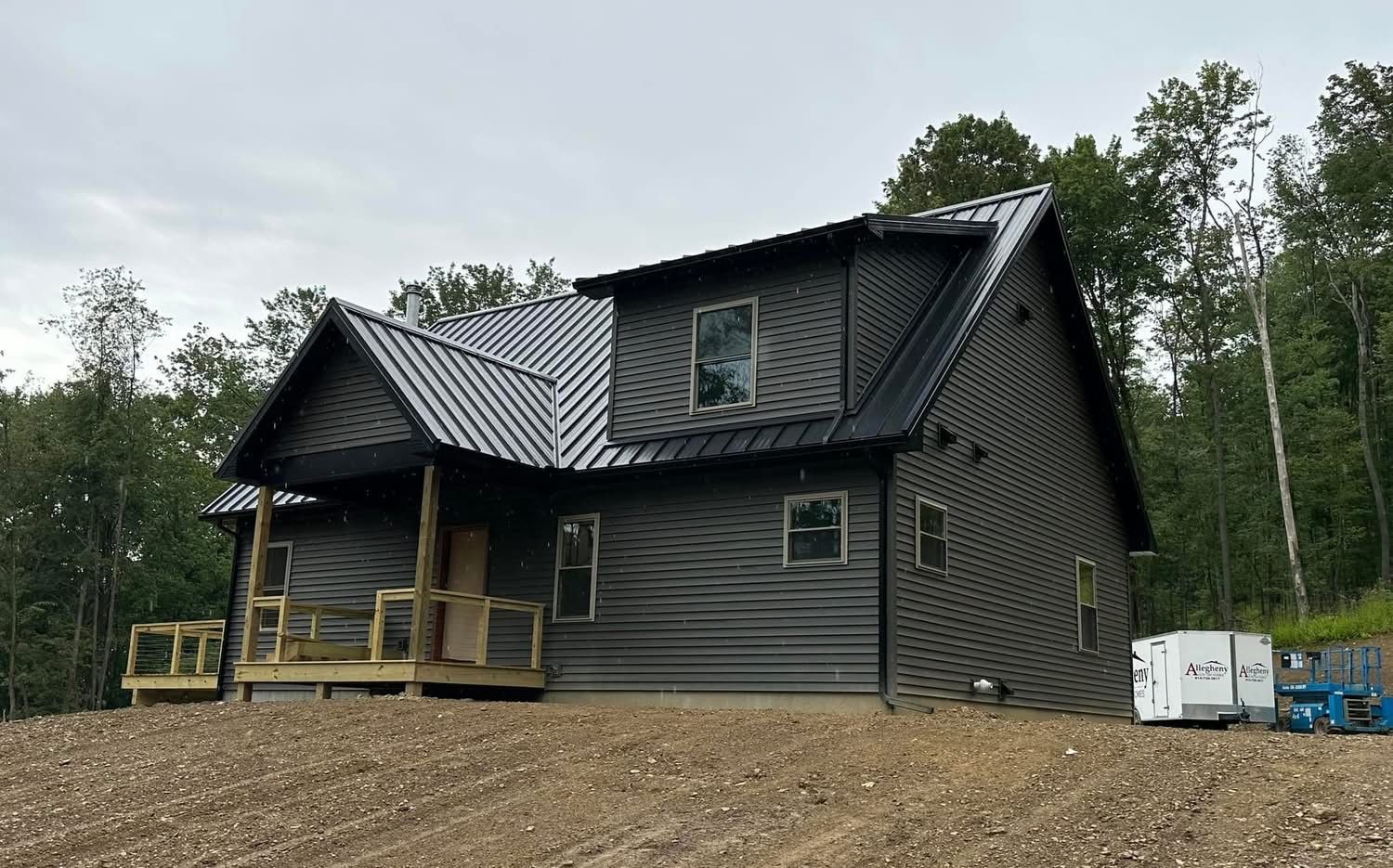 Dark gray cabin under construction with a metal roof, porch, and gravel yard, surrounded by trees
