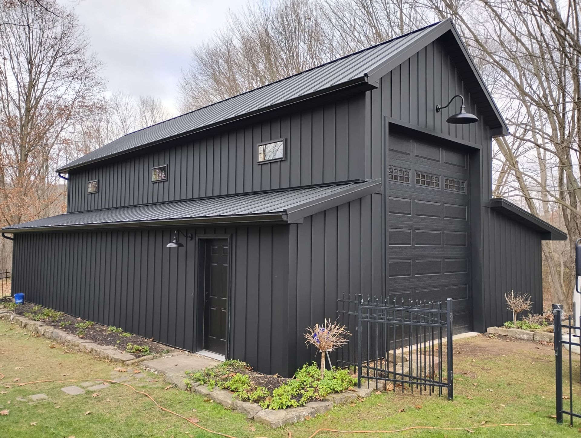 Black barn-style two-story building with corrugated siding, metal roof, and small fenced garden in a wooded area