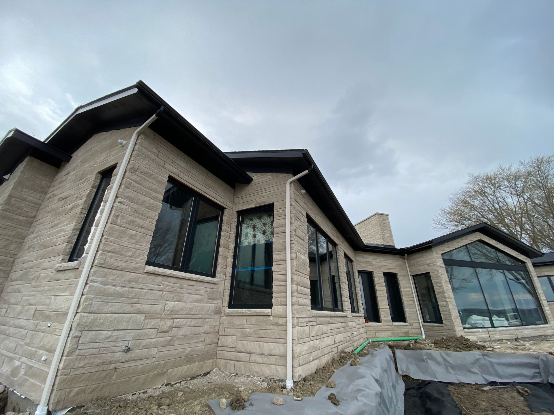 Stone house exterior with large windows and unfinished landscaping under a cloudy sky
