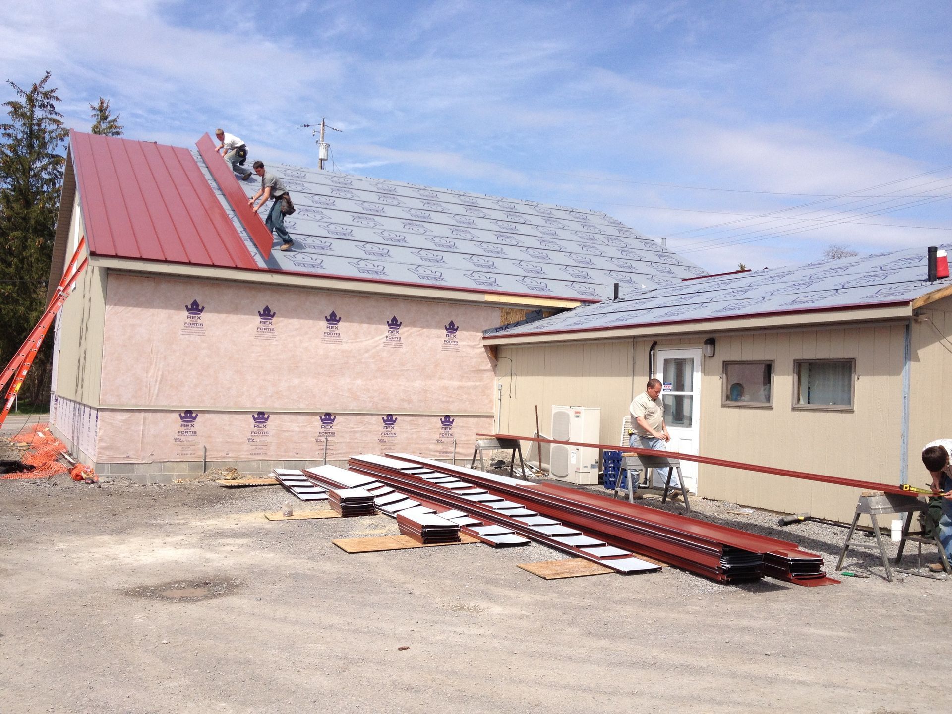 Construction workers on a roof beside stacked red metal beams and a beige building under a blue sky