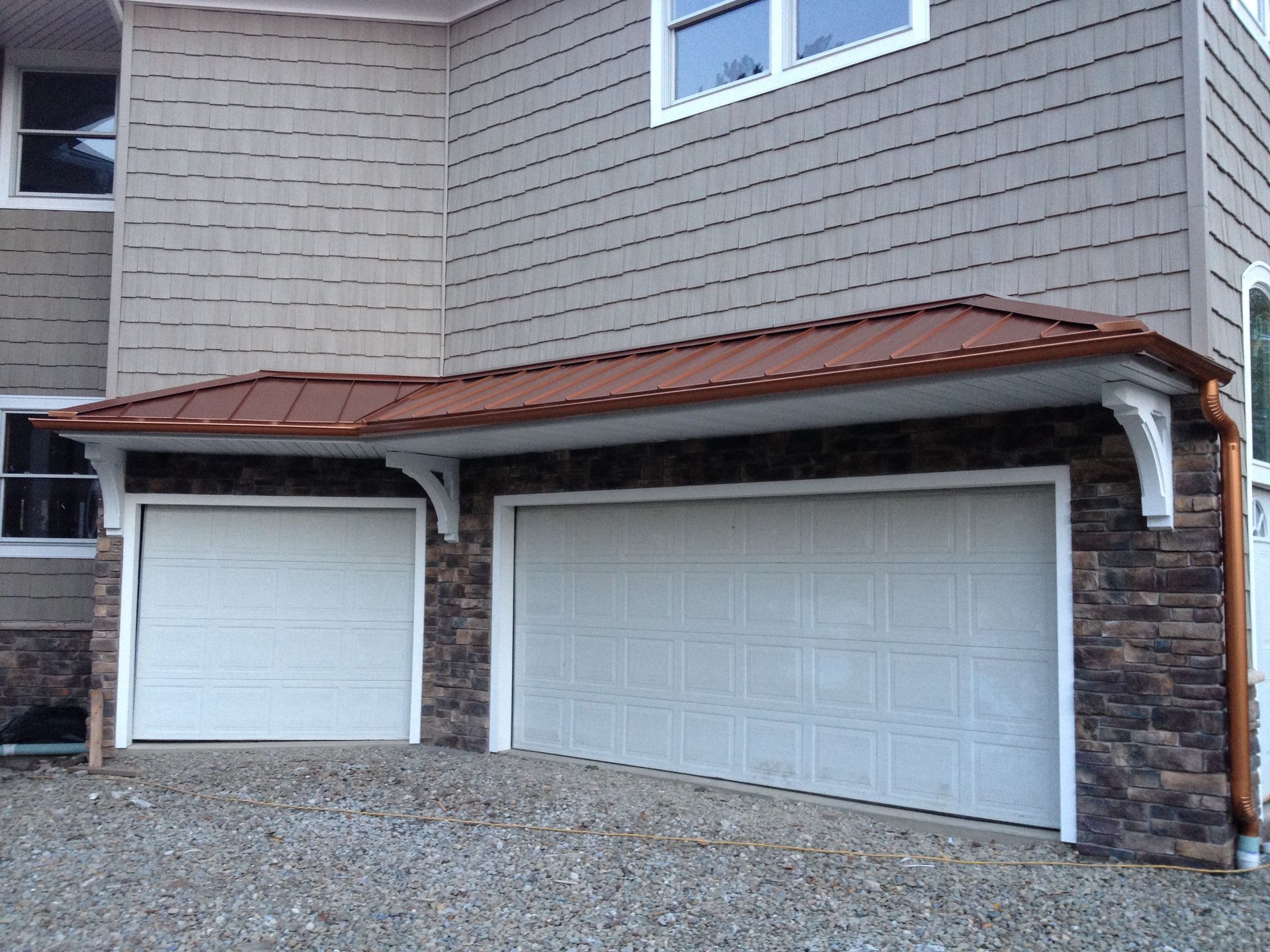 Two beige garage doors beneath a red metal awning on a gray stone-and-siding building