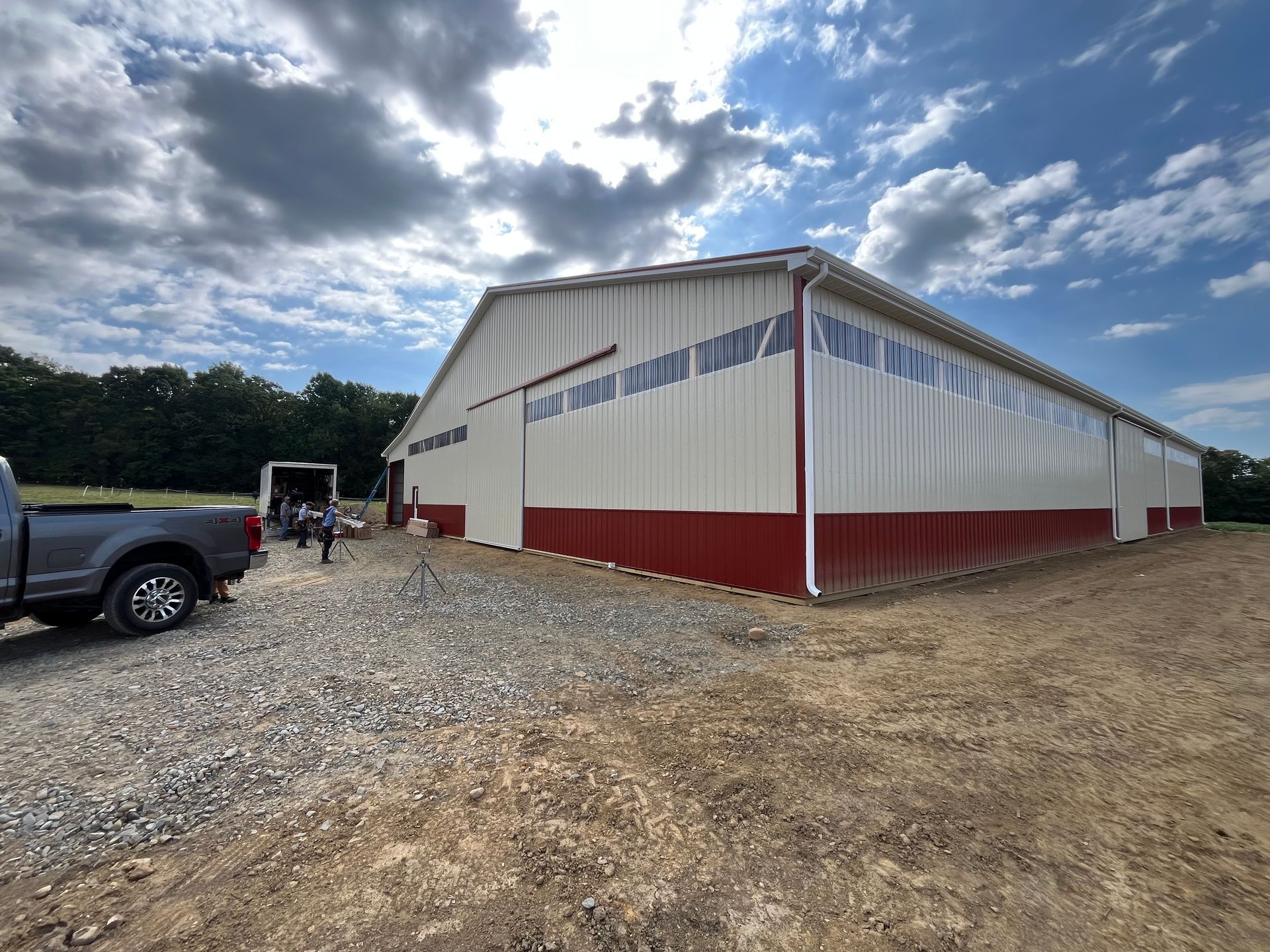 Gravel lot with a large white-and-red barn-style building under a cloudy sky, with vehicles parked nearby