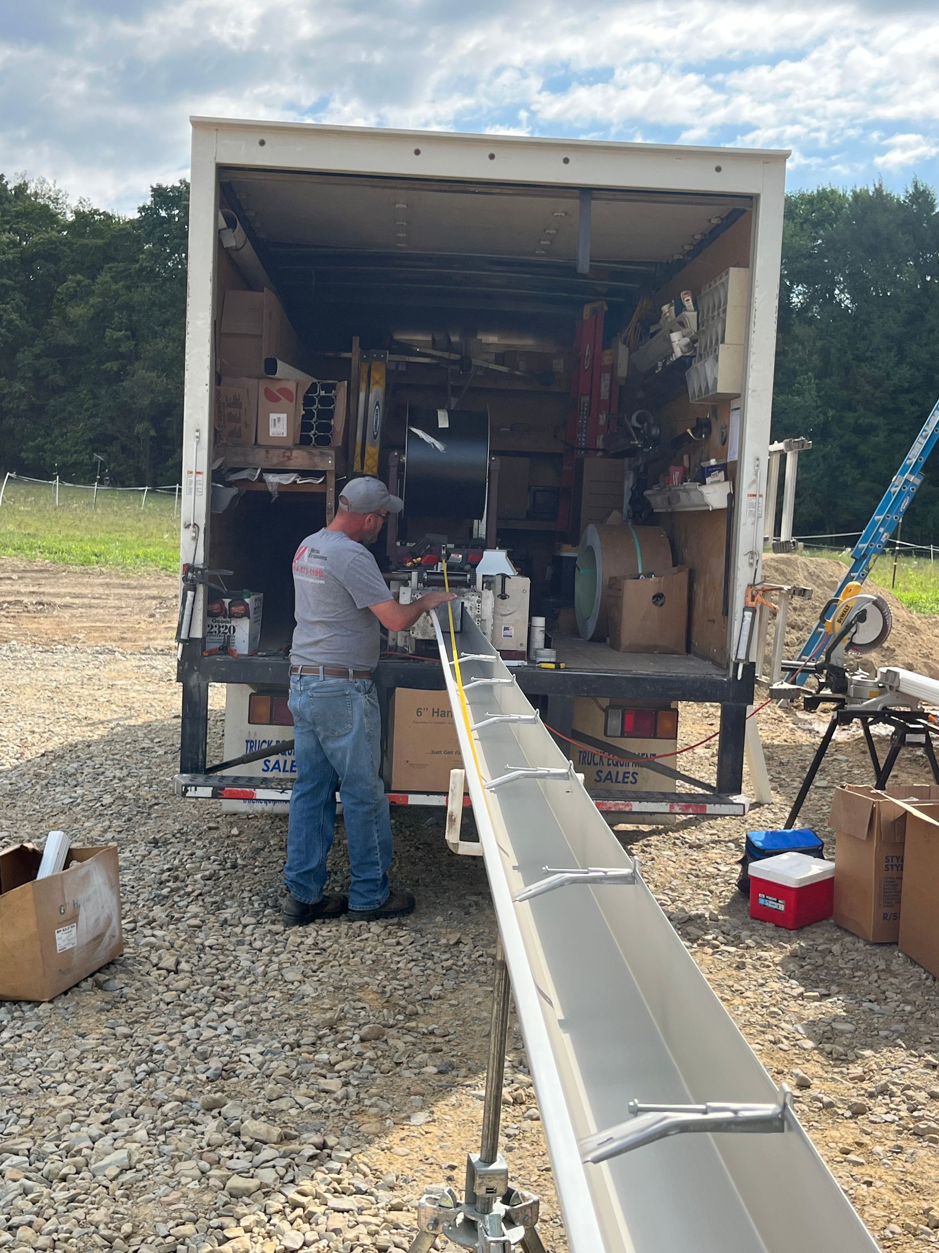 Man loading a long metal beam into a box truck in a gravel lot under cloudy skies