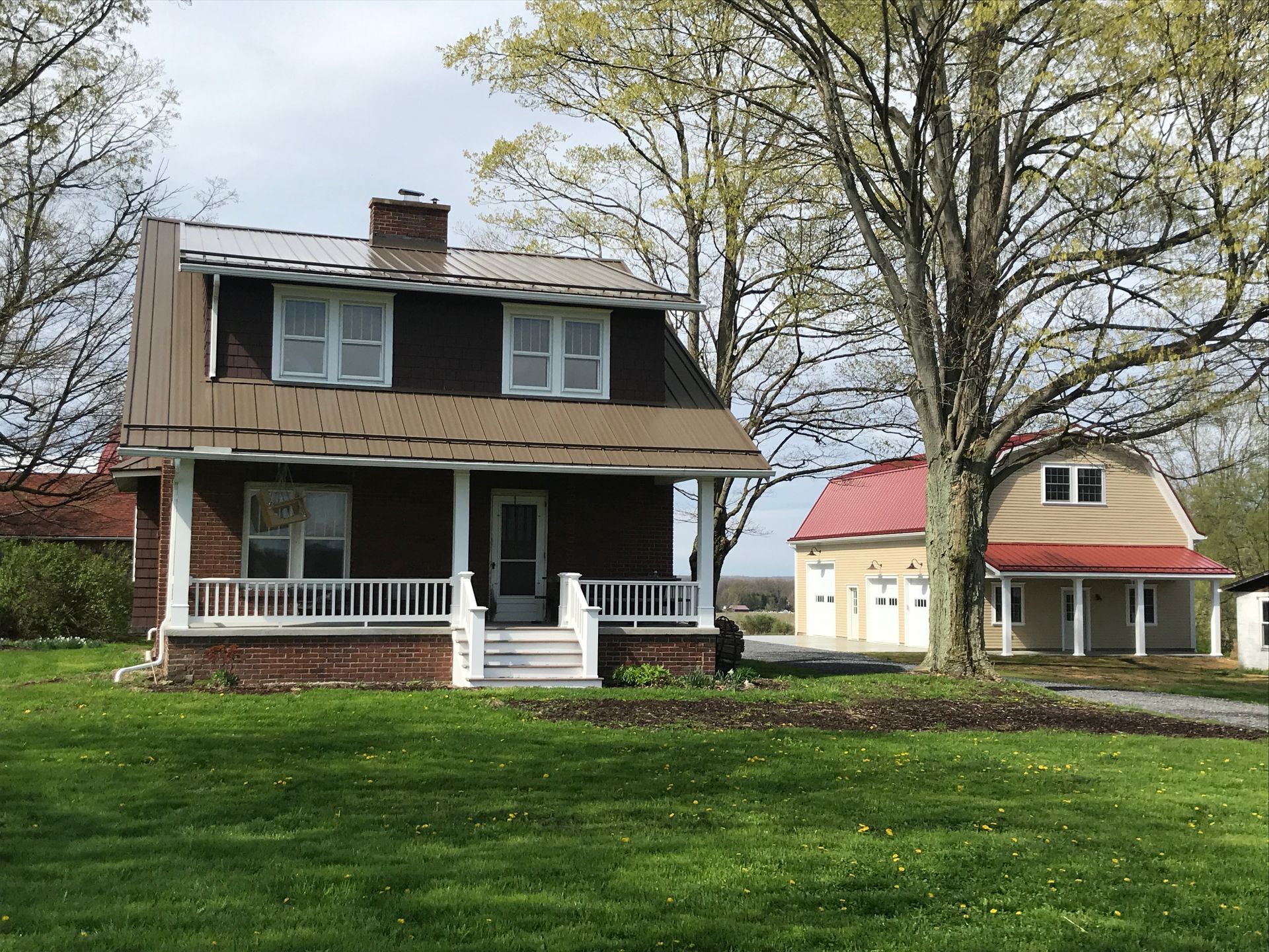 Two-story house with brown siding and porch on a green lawn, with a small yellow outbuilding and red-roofed gazebo nearby