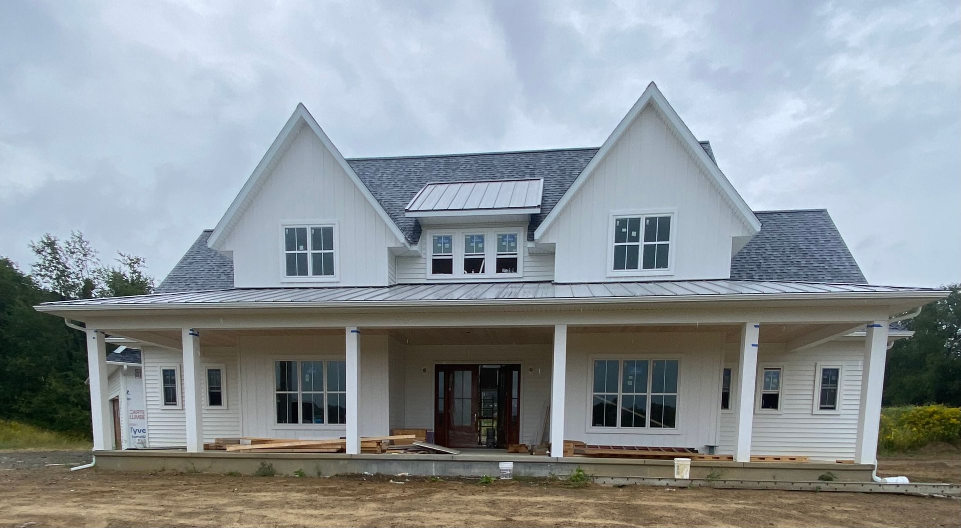 Large white farmhouse with gabled roof and wraparound porch under construction on a dirt lot.