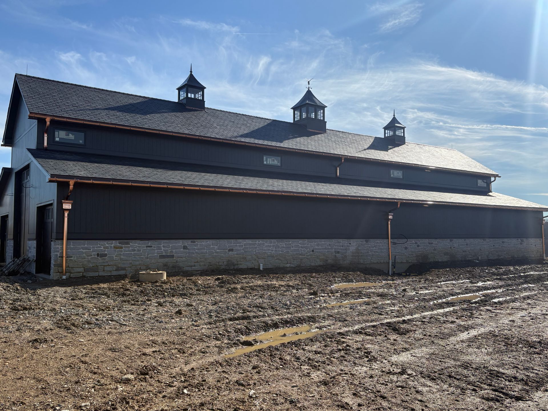 Large dark barn with cupolas in a muddy field under a bright blue sky